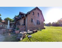 Backsteinbau mit Satteldach und dunkelgrünen Fensterläden in einer ländlichen Umgebung, umgeben von einer Rasenfläche. Auf der linken Seite stehen Gartenmöbel und Sonnenschirme auf einer Terrasse.