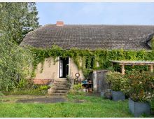 Ehemaliges Wohnhaus mit verwittertem Ziegeldach, dessen Fassade von üppigem Efeu und anderen Kletterpflanzen bedeckt ist. Vor dem Haus befinden sich eine alte Steintreppe, umliegendes Grün und ein kleiner hölzerner Anbau auf der rechten Seite.