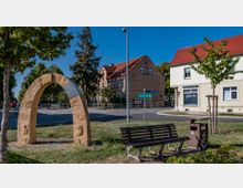 Kleiner Platz mit einer Steinbogen-Skulptur, die mit Wappen und Symbolen verziert ist. Im Hintergrund sind eine Straßenkreuzung, Bäume sowie Gebäude mit roten Ziegeldächern und heller Fassadengestaltung zu sehen.
