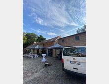 Ein rustikales Backsteingebäude mit Schrägdach auf einem gepflasterten Hof, links Pavillons mit dekorierten Stehtischen, rechts ein geparktes weißes Fahrzeug mit der Aufschrift "Kochschule Ben Perry". Im Hintergrund Bäume und ein blauer Himmel mit Wolken.