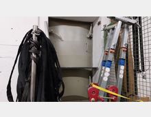 Blick in einen Lagerraum mit einer runden, metallenen Sicherheitstür an der Wand. Links hängen schwarze Gurte an einem Metallpfosten, rechts stehen mehrere zusammengefaltete Leitern an einem Maschendrahtzaun.