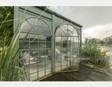 Grünes, gläsernes Gewächshaus mit großen Bogentüren und Sprossenfenstern, umgeben von Schilf und Vegetation auf einer Holzplattform, mit Blick auf einen See und bewölkten Himmel im Hintergrund.