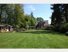 Ein rotes Backsteinhaus mit Satteldach und Balkon steht inmitten eines großen, gepflegten Gartens mit Rasenfläche, umgeben von Bäumen und dichter Vegetation. Links im Vordergrund befindet sich ein kleines rotes Gartenhäuschen mit weißer Tür und Fenstern.