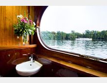 Innenraumansicht eines Bootes mit Holzverkleidung, einem runden Fenster mit Blick auf einen See und bewaldetes Ufer im Hintergrund. Neben dem Fenster befindet sich ein Waschbecken und eine Vase mit Blumen.