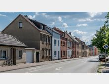 Reihenhäuser mit unterschiedlichen Fassaden aus Putz und Klinker stehen an einer ruhigen Straße, die von Gehwegen mit vereinzelten Fahrrädern und Bäumen gesäumt ist. Der Himmel ist leicht bewölkt, und einige geparkte Autos stehen am Straßenrand.