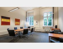 Konferenzraum mit einem ovalen Holztisch und acht schwarzen Stühlen, hellen Wänden, zwei großen Fenstern mit Blick auf grüne Bäume, abstrakter Wandkunst, einer Flipchart und einer kleinen Sitzecke mit Ledersesseln und einem kleinen Tisch.