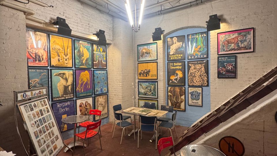 Kleiner Innenraum mit weiß gestrichenen Ziegelwänden, dekoriert mit bunten Plakaten, die Tiere und Zoothemen zeigen. Der Raum verfügt über Metalltische mit roten und blauen Stühlen, eine rötlich-braune Bodenfläche und eine auffällige Hängelampe unter der hohen Decke.