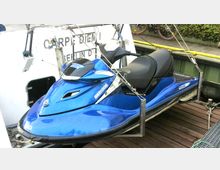 Ein blaues Jetboot (Modell Sea-Doo GTX) ist aufgehängt und seitlich an einem Boot befestigt. Im Hintergrund sind Teile des Bootes mit der Aufschrift „CARPE DIEM BERLIN“ sowie ein Uferbereich mit Fahrrädern zu sehen.