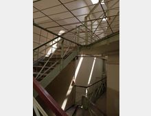 Innenansicht eines Treppenhauses in einem alten Gebäude mit Metallgeländern und grünen Stahlstreben. Sonnenlicht fällt durch ein Oberlicht und erzeugt helle Streifen auf den beigen Wänden und den Treppenstufen.