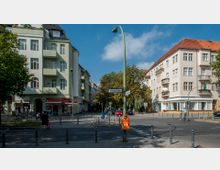 Straßenszene mit mehrstöckigen Wohnhäusern im Altbaustil, die helle Fassaden und Balkone haben. Eine Straßenkreuzung mit Verkehrszeichen, Gehwegen, Pollern und einer Straßenlaterne mit Straßenschildern „Bundesallee“ und „Trautenaustraße“ ist zu sehen, daneben Bäume, geparkte Autos und ein Fahrradfahrer.