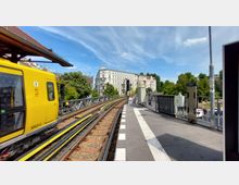 Eine oberirdische U-Bahn-Station mit einem gelben U-Bahn-Wagen auf den Schienen; im Hintergrund sind weiß verputzte Altbaugebäude sowie Bäume unter einem blauen Himmel zu sehen. Auf dem Bahnsteig befinden sich ein Signal und verschiedene technische Einrichtungen.