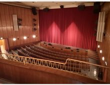 Innenraum eines mittelgroßen Theatersaals mit hölzerner Wandverkleidung, roten Vorhängen auf der Bühne und mehreren Sitzreihen mit braunen Polsterstühlen. An den Wänden sind Lampen und Lautsprecher angebracht, und eine goldfarbene Reling trennt den oberen Bereich vom Parterre.