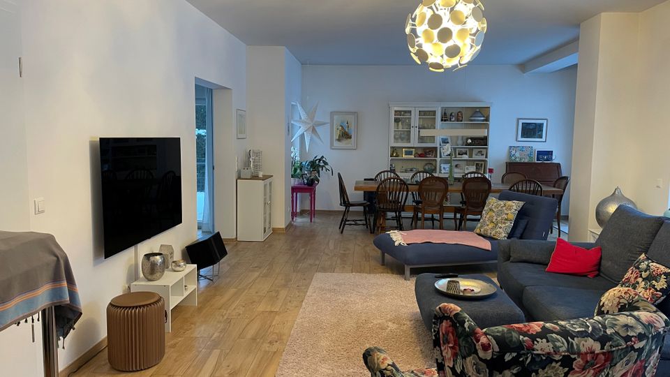 Offener Wohn- und Essbereich in einer Wohnung mit hellen Wänden und Holzboden, mit Sofaecke auf Teppich sowie einem an der Wand montierten Fernseher. Im Hintergrund steht ein großer Esstisch vor einem weißen Regal, darüber hängt eine runde Design-Deckenleuchte.