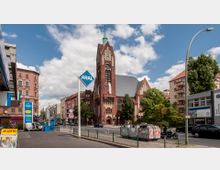 Straßenszene mit einer großen, markanten Backsteinkirche mit Spitzdach und grünem Turm im Zentrum. Im Vordergrund rechts stehen Container, links ist eine Aral-Tankstelle mit Werbeschildern, umgeben von mehrstöckigen Wohnhäusern und Bäumen.