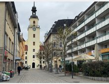 Eine gepflasterte Straße in einer Innenstadt mit einer gelben Kirchturmfassade und einer Uhr im Zentrum des Bildes. Auf beiden Seiten der Straße stehen Gebäude mit Geschäften, Balkonen und Cafés; vereinzelte Passanten und Fahrräder sind sichtbar.