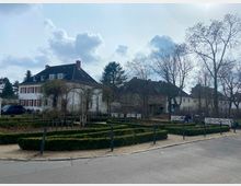 Eine von Hecken eingerahmte Gartenanlage mit symmetrischen Wegen im Vordergrund. Im Hintergrund stehen mehrere zweistöckige Wohnhäuser mit Satteldächern, umgeben von kahlen Bäumen und Sträuchern.