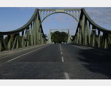 Grüne Stahlbrücke mit geschwungener Form und dekorativen Bögen, die über eine zweispurige Fahrbahn führt; im Hintergrund sind Bäume und ein blauer Himmel zu sehen.