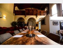 Innenraum eines umgestalteten historischen Gebäudes mit gewölbten Arkaden, verziertem hölzernem Balkon und hohen Fenstern. Im Vordergrund steht ein langer Holztisch mit Kerzenständer, umgeben von gepolsterten Stühlen, im Hintergrund befinden sich Bücherregale und eine gemütliche Sitzecke.