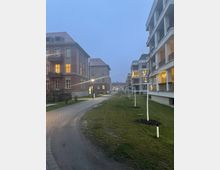 Wohnsiedlung mit mehrstöckigen Gebäuden, links aus rotem Backstein mit traditionellen Balkonen und rechts moderner Architektur mit offenen Balkonen. Dazwischen verläuft ein beleuchteter Gehweg, gesäumt von jungen Bäumen und gepflegten Grünflächen, bei dämmernder Abendstimmung.