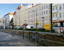Straßenszene mit mehrstöckigen Wohnhäusern in hellen Farben, einer Straßenbahnhaltestelle mit Wartehäuschen und parkenden Autos. Im Vordergrund sind Fahrradständer, Straßenbahnschienen und ein DHL-Lieferwagen zu sehen.