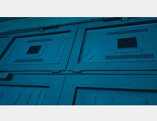 Nahaufnahme von einer metallischen Wand mit rechteckigen Platten, Luftschlitzen und wabenförmigen Belüftungsgittern. Die Oberfläche ist blau beleuchtet, und die Konstruktion weist industrielle oder technische Merkmale auf.