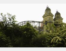 Steinernes Brückenportal mit verzierten Kuppeln und Metallornamenten, im Hintergrund eine Stahlbogenbrücke. Im Vordergrund dichte grüne Vegetation mit blühenden Sträuchern.