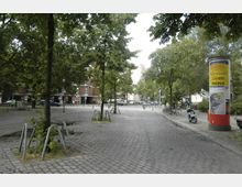 Städtischer Platz mit Kopfsteinpflaster und Bäumen, umgeben von Wohngebäuden. Im Vordergrund befinden sich Fahrradständer und eine Litfaßsäule mit Werbeplakaten, im Hintergrund parkende Autos und Fahrräder.