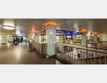 Unterirdischer Bahnhof mit Säulen, Leuchtstreifen und Schildern, die auf verschiedene Zuglinien und Ziele wie "Wannsee" und "Blankenfelde" hinweisen. Es sind Fahrgäste mit Gepäck sowie Treppen und Aufgänge sichtbar, die zu verschiedenen Ebenen führen.