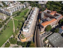 Luftaufnahme eines städtischen Gebiets mit einem Park auf der linken Seite, einem markanten Gebäudekomplex mit begrünten Dächern und einer angrenzenden Bahnstrecke. Rechts daneben befinden sich ein rotes Backsteingebäude und mehrere Wohnhäuser, eingerahmt von Straßen und Grünflächen.