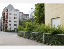 Mehrstöckige Wohngebäude in einer städtischen Umgebung, umgeben von Bäumen und Büschen mit einem Metallzaun. Im Hintergrund ist eine weiße Hauswand mit einem gemalten Regenbogen zu sehen.