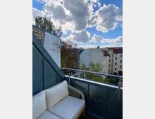 Teilansicht eines kleinen Balkons einer Wohnung mit grauem Geländer, das mit Lichterketten dekoriert ist. Im Vordergrund steht eine Sitzbank mit weißen Polstern, dahinter ist der Blick auf Nachbargebäude, Bäume und einen Himmel mit vielen Wolken zu sehen.