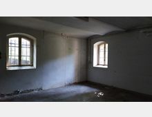 Ein leerer, ungepflegter Raum mit weiß gestrichenen Wänden und Betonboden. Zwei bogenförmige Fenster mit Gitterstäben lassen diffuses Tageslicht hinein.