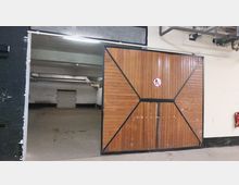 Holztor mit diagonal verlaufenden Metallverstärkungen, teilweise geöffnet, führt in einen leeren, betonierten Innenraum mit weißen Wänden und freiliegenden Rohren an der Decke. An der Tür befindet sich ein Verbotsschild, und rechts hängt ein Feuerlöscher an der Wand.