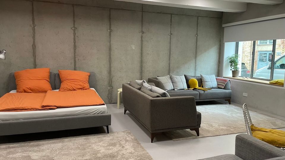 Industrial-style studio room with exposed concrete walls and ceiling beams, furnished with a low bed with orange bedding, several gray sofas, and light rugs on a smooth floor. A large window with a roller blind sits along one wall, with a potted plant on the sill and street views outside.