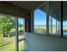 Ein Innenraum mit großen Fenstern, die einen Ausblick auf einen See, Schilf und grüne Wiesen bieten. Die Wände bestehen teils aus unverputztem Ziegelmauerwerk, Holzverkleidung an der Decke und einer Überdachung im Außenbereich wird sichtbar.