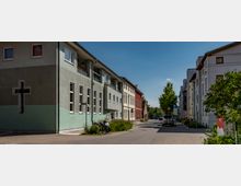 Straßenszene mit mehrstöckigen Wohngebäuden im Vordergrund, darunter ein grünes Gebäude mit einem Kreuzfenster an der Seite. Fußwege und vereinzelte Bäume säumen die Straße, die in einen ruhigen, sonnigen Bereich führt.