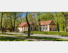 Low, historic buildings with faded stucco walls and red tile roofs sit along a quiet road in a leafy park setting. Tall trees surround the structures, including one section with large multi-pane windows and timber framing on the gable.