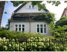 Wohnhaus mit heller Putzfassade und Satteldach, umgeben von üppigem, grünem Garten mit Hortensien und Metallzaun im Vordergrund. Ein Balkon im Obergeschoss ist mit Pflanzen und Kletterpflanzen dekoriert.