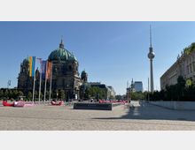 Weitläufiger Platz in Berlin mit Blick auf den Berliner Dom im linken Bildbereich, flankiert von Fahnenmasten und Sitzgelegenheiten. Im Hintergrund ist der Fernsehturm sowie moderne und historische Gebäude zu sehen, während rechts ein klassizistisches Gebäude mit Säulen und Fassadenverzierungen steht.