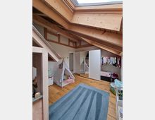 Dachgeschoss-Kinderzimmer mit sichtbaren Holzbalken, einem großen Dachfenster und einem Boden aus hellem Holz. Im Raum stehen eine weiße Kleiderschrankwand mit integriertem Spiegel, ein kleines Zelt mit Decken und Kissen sowie eine Spielzeugkiste neben einer Garderobenstange mit Kinderkleidung. Ein blauer Teppich bedeckt teilweise den Boden.