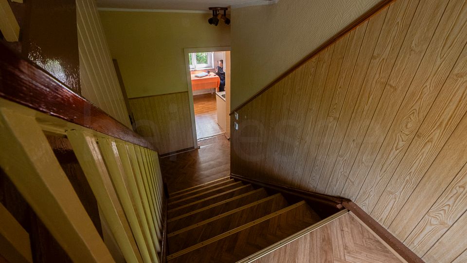 Blick in ein enges Treppenhaus mit Holzstufen, dunkelbraunem Handlauf und holzvertäfelter Wandverkleidung. Unten führt ein Flur zu einer offenen Tür in einen hellen Raum mit Fenster und Tisch.
