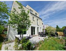 Modernes Wohnhaus mit hellbeiger Fassade, großen Fensterfronten und einer gepflasterten Terrasse. Auf der Terrasse stehen eine Sitzecke mit Sofa und ein Esstisch mit Stühlen, umgeben von einem gepflegten Garten mit Büschen, Blumen und einem kleinen Baum.