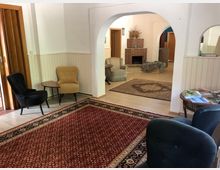 Ein heller Raum mit Sitzbereichen, darunter schwarze, beige und grüne Polsterstühle, ein roter Orientteppich und ein kleiner Holztisch. Im Hintergrund ist ein gemauerter Kamin mit Dekorationen und ein beige gemusterter Teppich sichtbar. Weiße Wände und Holzvertäfelungen bestimmen das Ambiente.