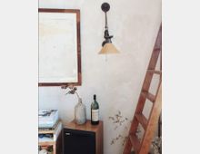 Ein Innenraum mit einer hellen, strukturierten Wand, an der eine retro-inspirierte Wandleuchte angebracht ist. Links steht ein Holzregal mit Büchern, daneben ein kleiner Holzschrank mit einer Glasvase, einer Weinflasche und einem Lautsprecher; rechts ist eine Holzleiter sichtbar.