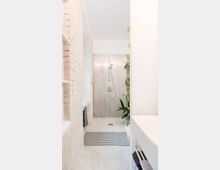 Helles, modernes Badezimmer mit begehbarer Dusche, weißen Fliesen im Mosaikstil und einer Rückwand mit vertikaler Struktur. Links ist eine gemauerte Wand in Weiß und eine eingelassene Badewanne erkennbar, rechts stehen grüne Pflanzen als Dekoration.