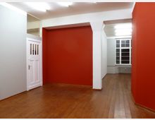 Ein leerer Innenraum mit Holzboden, weißen Wänden und einem roten Akzentbereich, einer weißen Tür links, und einem großen Fenster im Hintergrund, das Licht in den Raum lässt.