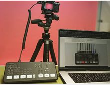 Ein Kamerasetup mit Stativ und angeschlossener Kamera vor einem roten Hintergrund, daneben ein Laptop mit geöffneter Webseite und ein ATEM Mini Videomischer auf einem Tisch.