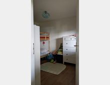 Kinderzimmer mit Hochbett aus Holz auf der linken Seite, einer weißen Kommode auf der rechten Seite und einem kleinen Teppich mit Straßenmotiv in der Mitte. Der Raum besitzt Holzboden und eine Deckenleuchte mit blauer Glasschirm.