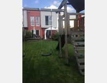 Spielplatz mit einer Holz-Kletterstruktur, Schaukeln und einer Rutsche in einem Garten. Im Hintergrund sind ein modernes Wohnhaus mit roten und weißen Fassaden sowie eine Terrasse mit Sonnenschirmen sichtbar.