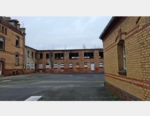 Innenhof eines verlassenen Backstein-Industriekomplexes mit mehrstöckigen Gebäudefassaden, teils vernagelten oder zerstörten Fenstern und dunklen Öffnungen. Der asphaltierte Hof ist weitgehend leer, umgeben von gelb-rotem Mauerwerk unter grauem Himmel.